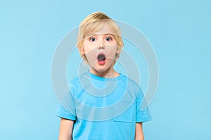 Portrait of a shocked cute little boy with blond hair on pastel blue background. Surprised preschooler studio shot.