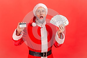 Portrait of shocked elderly man with gray beard wearing santa claus costume holding fan of dollars