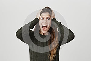 Portrait of shocked european woman screaming with popped eyes holding hands on head in dark-green sweater, isolated over