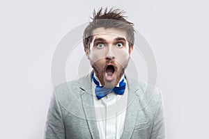 Portrait of shocked handsome bearded man in casual grey suit and blue bow tie standing with big eyes and open mouth and looking at
