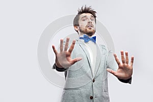 Portrait of shocked handsome bearded man in casual grey suit and blue bow tie standing with blocking hands and looking at camera