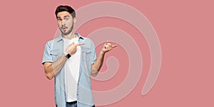 Portrait of shocked handsome bearded young man in blue casual shirt standing, looking at camera with surprised face and pointing