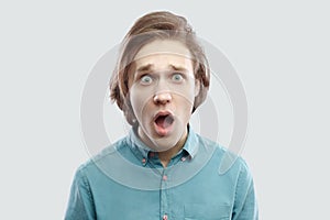 Portrait of shocked handsome long haired blonde young man in blue shirt standing with big eyes, open mouth, surprised face looking