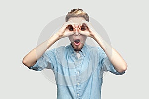 Portrait of shocked handsome young man in light blue shirt standing with hands on eyes binoculars gesture and looking at camera