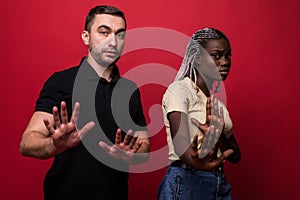 Portrait of shocked interracial couple: African woman and Caucasian man standing together against red background, covering face,