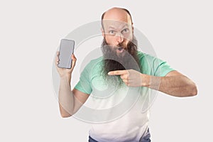 Portrait of shocked middle aged bald bearded man standing with mobile smart phone pointing at empty screen and looking with