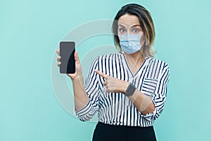 Portrait of shocked middle aged woman with surgical medical mask standing and showing mobile smart phone display with big eyes