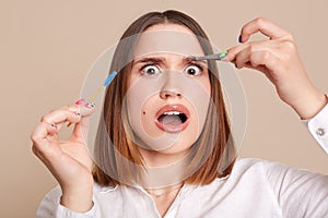 Portrait of shocked scared female in beauty salon holding eyebrow brush and tweezers, looking at camera with big eyes, being