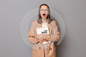 Portrait of shocked scared woman wearing beige jacket standing with organizer and calculator in hands isolated over gray
