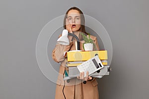 Portrait of shocked scared woman wearing stylish beige jacket holding office stuff posing isolated over gray background, keeps