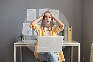 Portrait of shocked scared woman wearing yellow jacket sitting at workplace and working on laptop, looking at camera with open