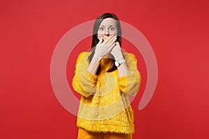 Portrait of shocked scared young woman in yellow fur sweater covering mouth with hands isolated on bright red wall