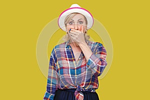 Portrait of shocked stylish mature woman in casual style with hat standing, covering her mouth and looking at camera with