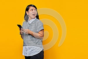 Portrait of shocked and surprised young Asian woman holding mobile phone and looking sideways on yellow background