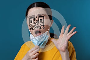 Portrait of a shocked woman with a QR code instead of eyes and nose, removing protective mask from her face. Blue