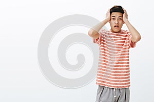 Portrait of shocked young asian guy in stupor pressing hands to head open mouth from amazement, being speechless photo