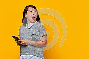 Portrait of shocked young Asian woman holding mobile phone and looking aside on yellow background