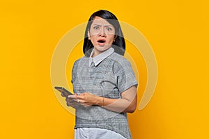 Portrait of shocked young Asian woman holding mobile phone on yellow background