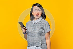 Portrait of shocked young Asian woman holding mobile phone on yellow background