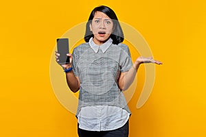 Portrait of Shocked young Asian woman showing smartphone blank screen and pointing to empty space with palms over yellow