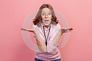 Portrait of shocked young brunette teenager girl in eyeglasses throwing hands up and shrugging shoulders, looking with big eyes