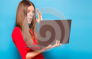 Portrait of shocked young girl holding laptop computer isolated over blue background
