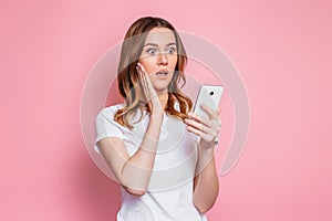 Portrait of a shocked young girl looking at phone and reads a messages or news  over pink background. Surprised woman in a