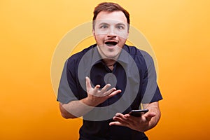 Portrait of shocked young man in blue shirt holding mobile phone isolated on yellow background