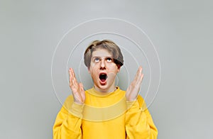 Portrait of shocked young man in bright clothes isolated on gray background, looking up with open mouth in surprise and arms