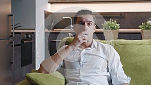 Portrait of shocked young man covers his face with his hand in fear while sits on sofa at home. Astonished man scared by