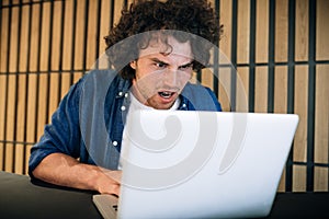 Portrait of shocked young man with curly hair working on laptop surfing on Internet, reading the news in the morning. Angry male