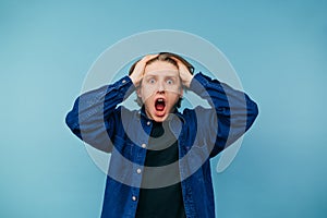 Portrait of a shocked young man in a shirt on a blue background, holding his head in fear and looking into the camera with his