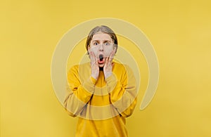 Portrait of a shocked young man on a yellow background, holding hands on his cheeks and looking scared at the camera