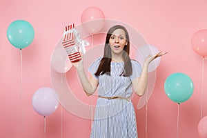 Portrait of shocked young woman in blue dress spreading hands holding red box with gift present on pink background with