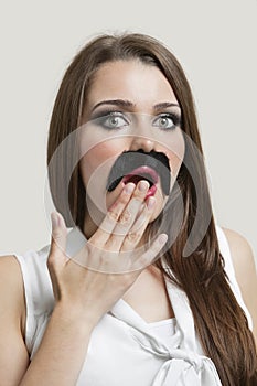 Portrait of shocked young woman with fake moustache over gray background