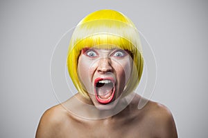 Portrait of shocked young woman with freckles, red makeup and yellow wig looking at camera with unbelievable face and screaming
