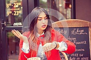 Portrait of shocked young woman holding hand with wrist watch and looking at camera isolated on coffee shop outside terrace