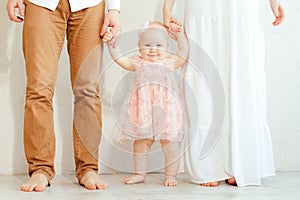 Portrait of shoeless mother and father holding hands of smiling baby girl in pink holiday dress on white background