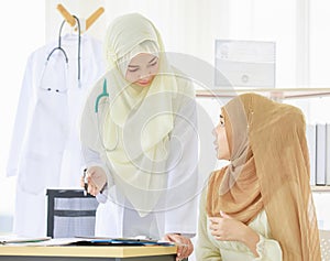 Portrait shot of adult Muslim doctor wearing hijab sitting at the table, talking to a patient while using a digital tablet.