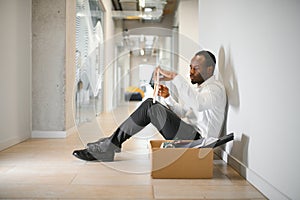 Portrait shot of African American sad businessman in suit and tie sitting with box of stuff. Male office worker lost job