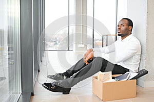 Portrait shot of African American sad businessman in suit and tie sitting with box of stuff. Male office worker lost job