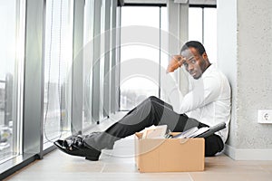 Portrait shot of African American sad businessman in suit and tie sitting with box of stuff. Male office worker lost job