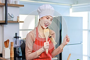 Portrait shot of Asian young beautiful female chef housewife wears white tall cook hat and apron standing smiling posing with