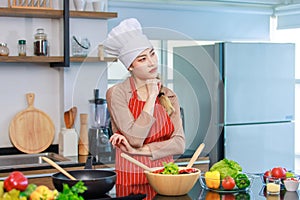 Portrait shot of Asian young beautiful female chef housewife wears white tall cook hat and apron standing smiling posing thinking