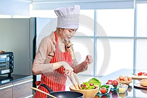 Portrait shot of Asian young female chef housewife wears white tall cook hat and apron standing smiling holding wooden spoon fork