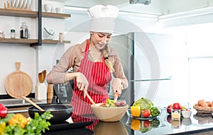 Portrait shot of Asian young female chef housewife wears white tall cook hat and apron standing smiling holding wooden spoon fork