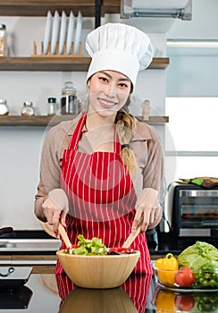 Portrait shot of Asian young female chef housewife wears white tall cook hat and apron standing smiling holding wooden spoon fork