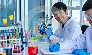 Portrait shot of Asian young male scientist in white lab coat and rubber gloves sitting smiling using microscope in laboratory