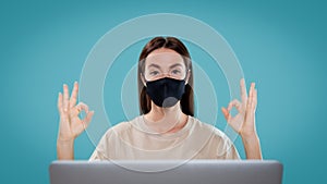 A portrait shot of cute brunette female works isolated with her computer via freelance, she wear mask and shows ok signs