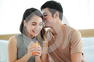 Portrait shot of cute smiling young Asian lover couple sitting on a bed together at home in the morning. Wife holding and drinking